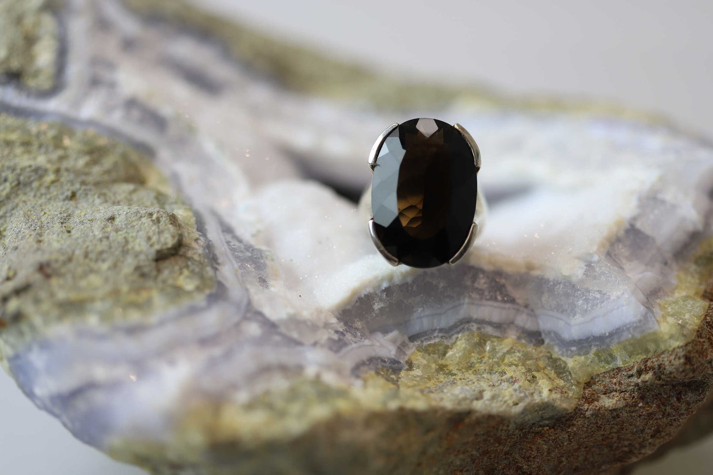 Kristallwerk präsentiert einen leuchtenden facettierten und ovalen Rauchquarz Ring aus Silber massiv auf einer Chalcedongeode..