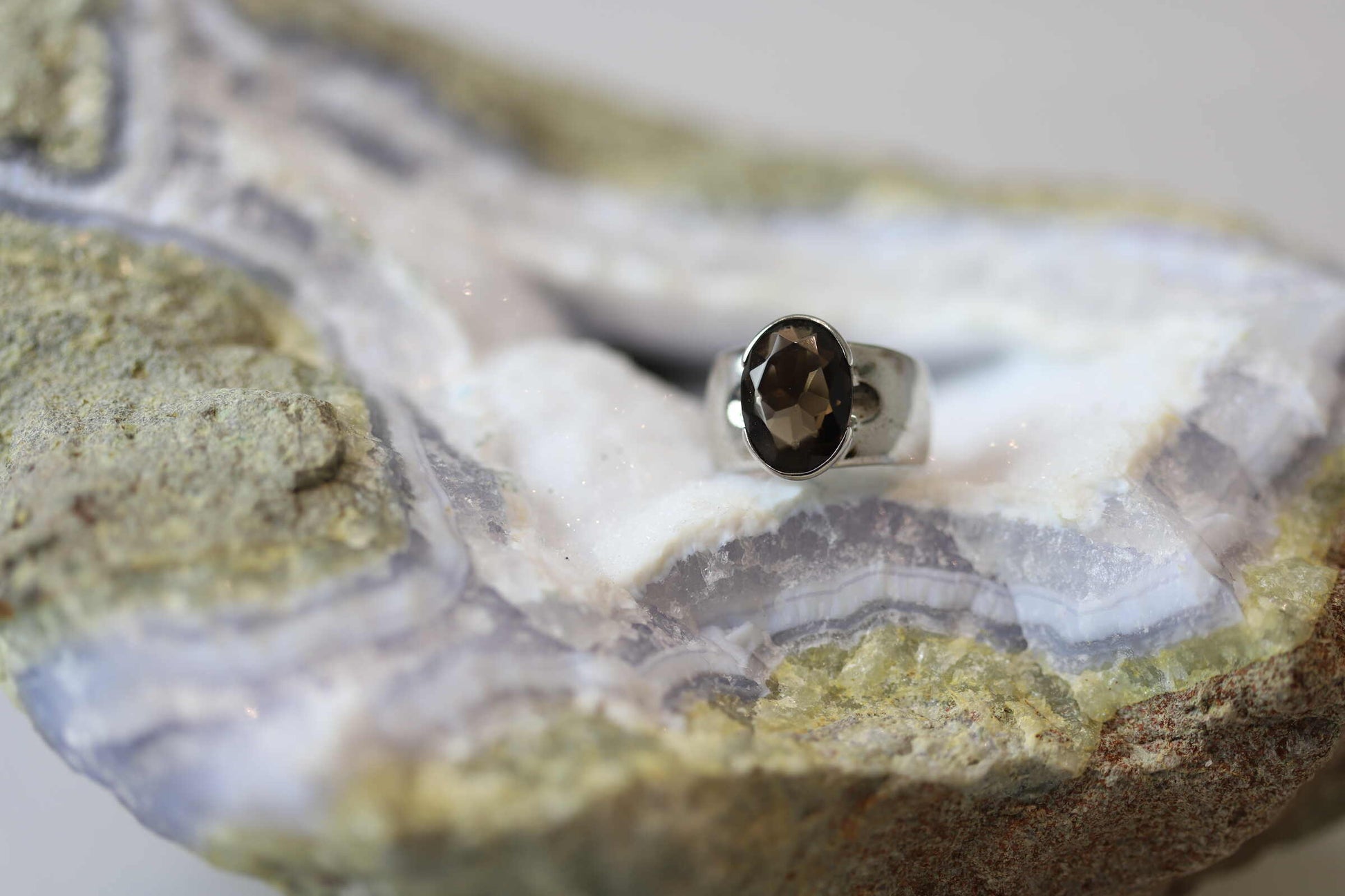 Kristallwerk präsentiert einen leuchtenden facettierten und ovalen Rauchquarz Ring aus Silber massiv auf einer Chalcedongeode..