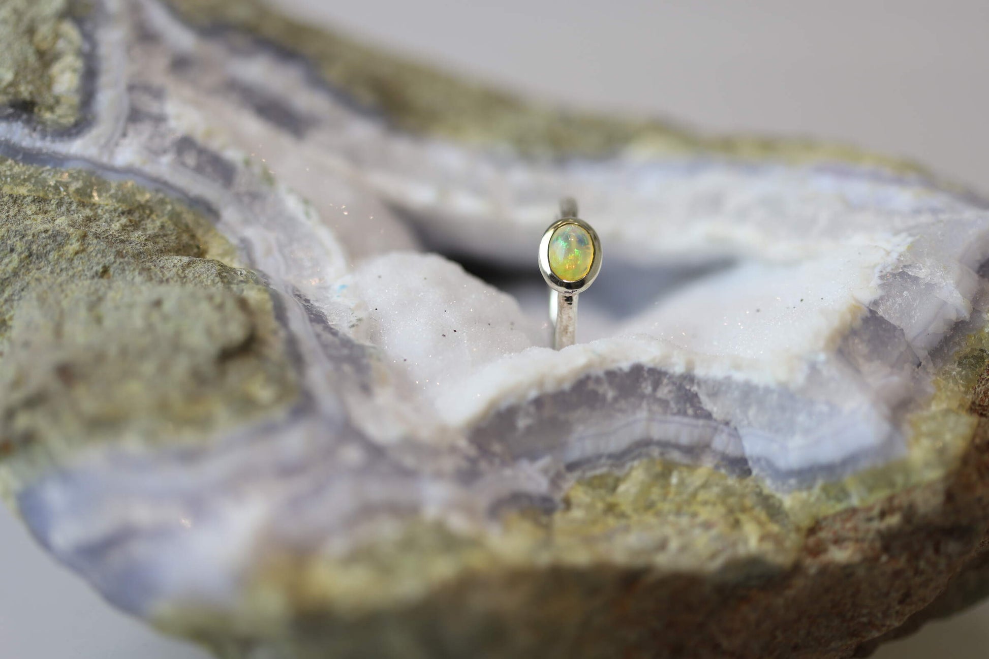 Kristallwerk präsentiert einen leuchtenden, irisierenden und ovalen Opal Ring aus Silber auf einer Chalcedongeode..