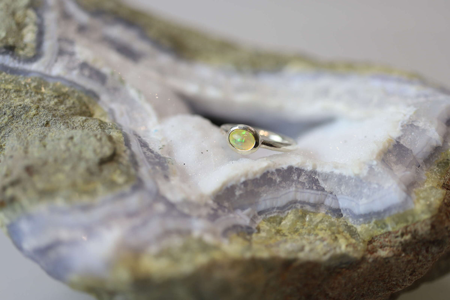 Kristallwerk präsentiert einen leuchtenden, irisierenden und ovalen Opal Ring aus Silber auf einer Chalcedongeode..