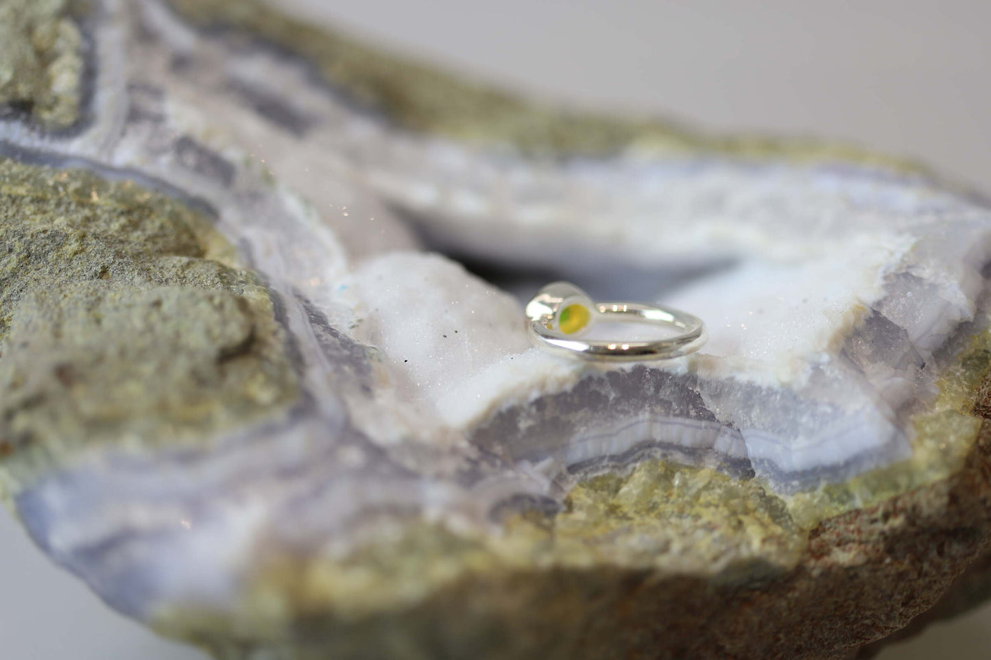 Kristallwerk präsentiert einen leuchtenden, irisierenden und ovalen Opal Ring aus Silber auf einer Chalcedongeode..