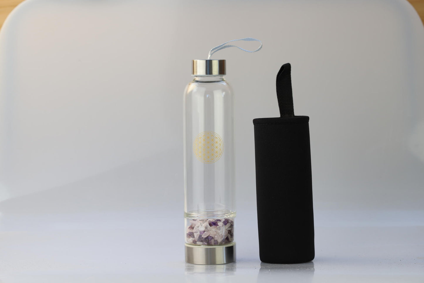 Kristallwerk präsentiert: Trinkflasche mit aufgedruckter Blume des Lebens mit Chips aus Amethyst, Bergkristall und Rosenquarz auf weiss glänzendem Untergrund.