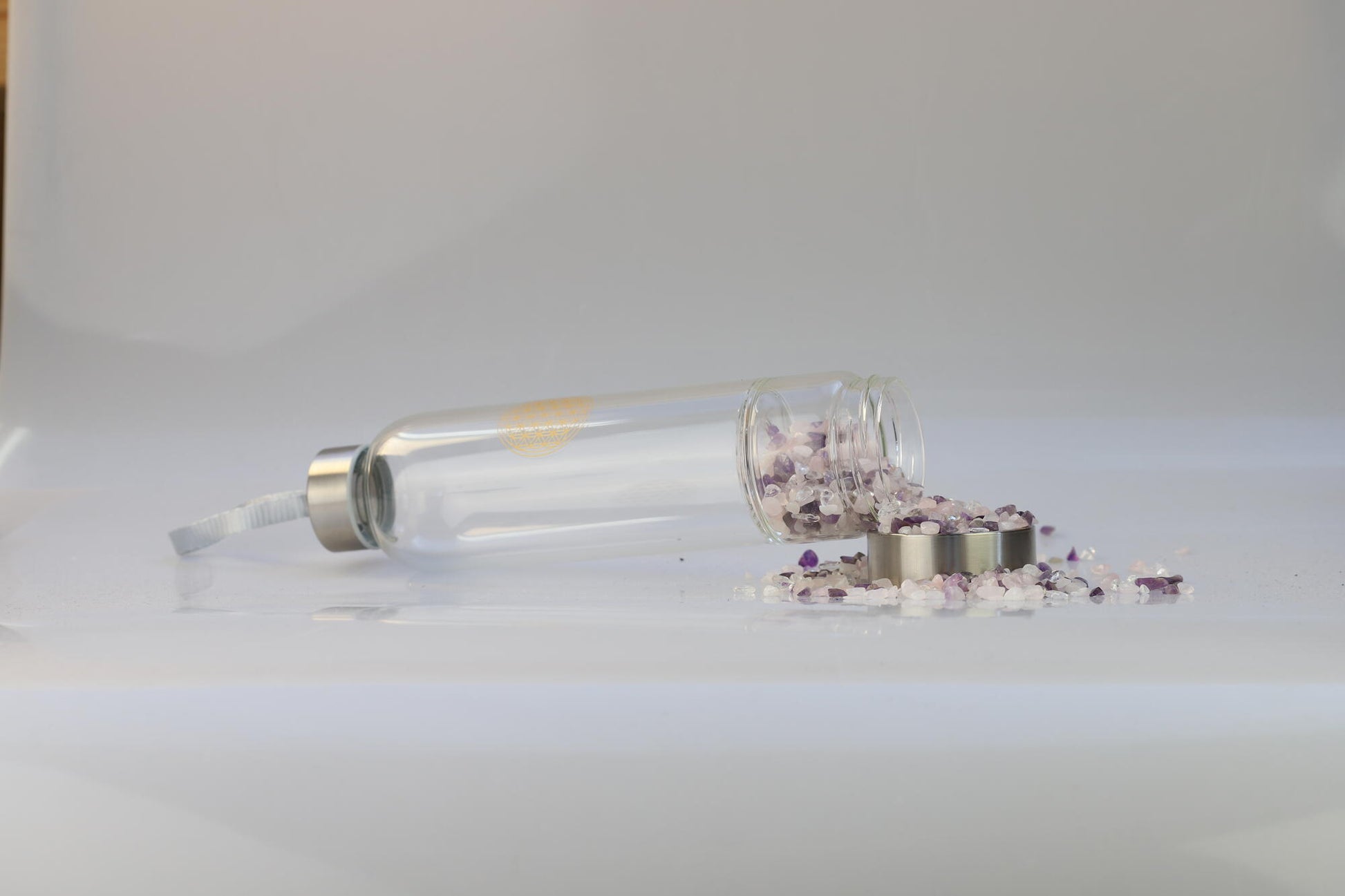 Kristallwerk präsentiert: Trinkflasche mit aufgedruckter Blume des Lebens mit Chips aus Amethyst, Bergkristall und Rosenquarz auf weiss glänzendem Untergrund.