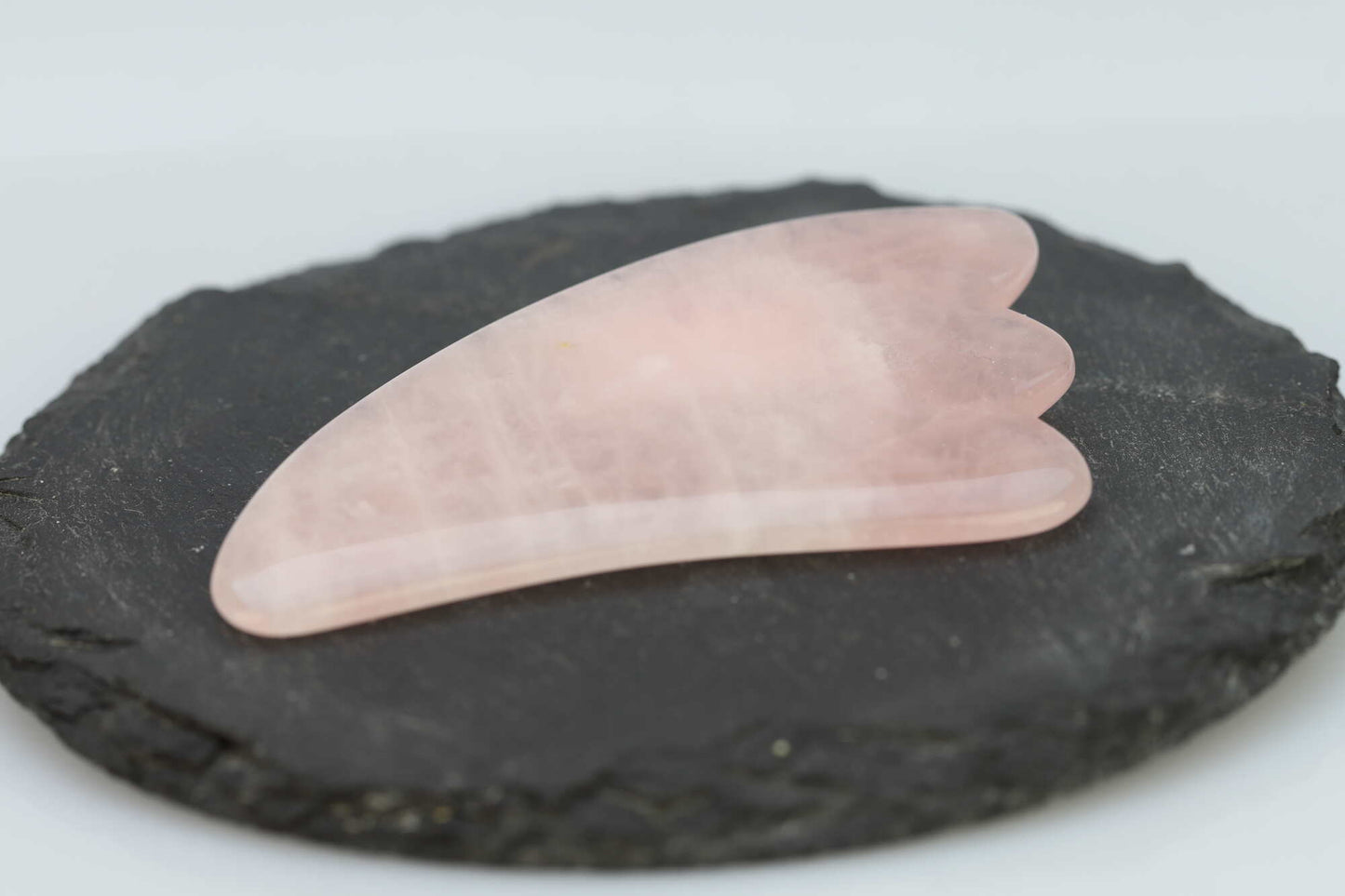 Kristallwerk präsentiert: Geschliffene Schätze Gua Sha Massagestück aus Rosenquarz auf einer Granitplatte.